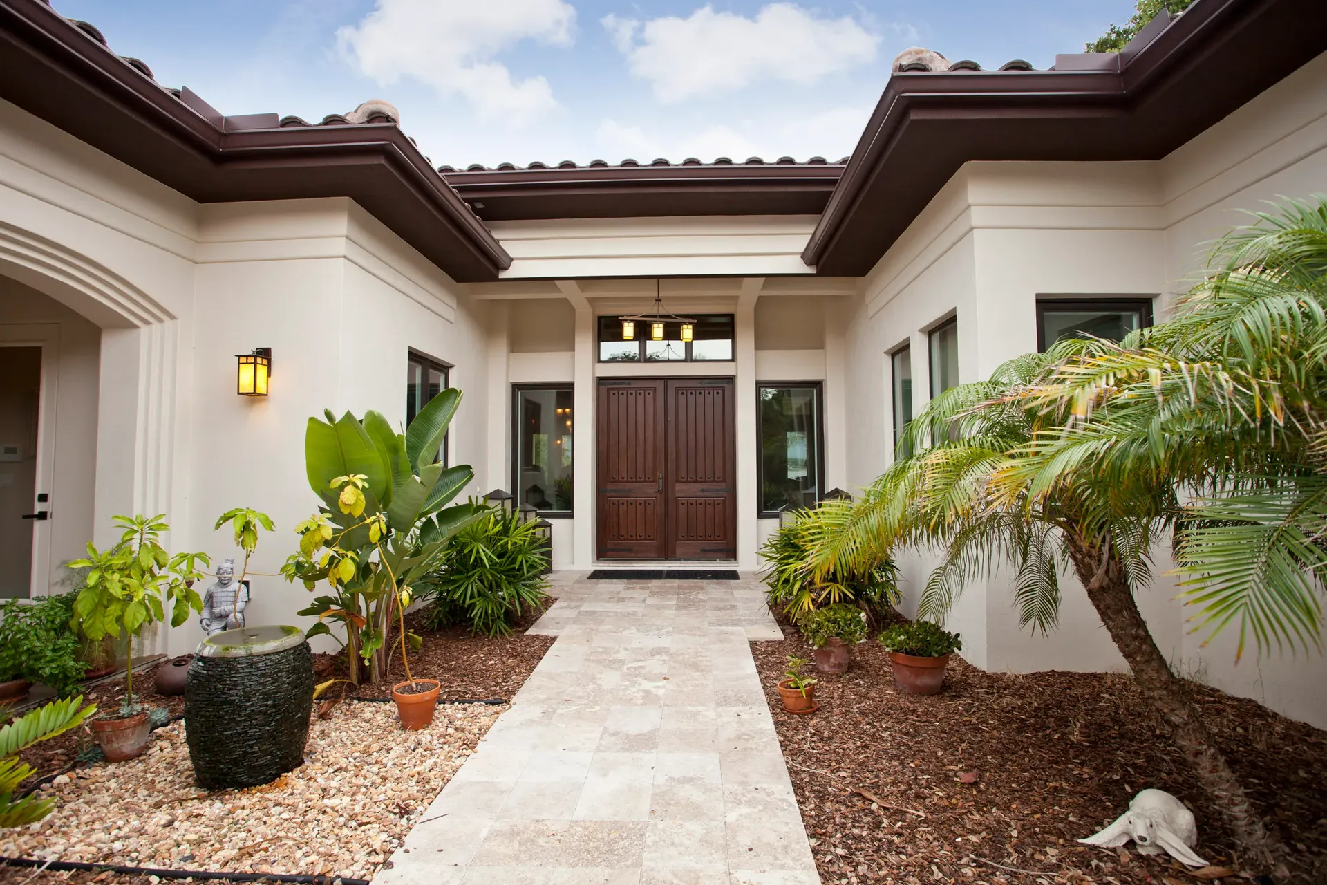 A Mediterranean-style home entrance with double wooden doors, lush greenery and stone path by house designer Ben Butera.